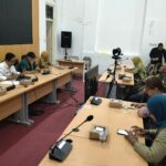 People sit at a long conference table with microphones and boxed favors during a meeting; a camera on a tripod is set up in the center of the room.
