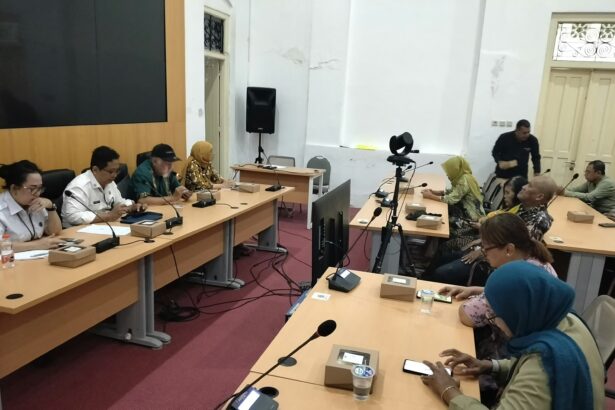 People sit at a long conference table with microphones and boxed favors during a meeting; a camera on a tripod is set up in the center of the room.