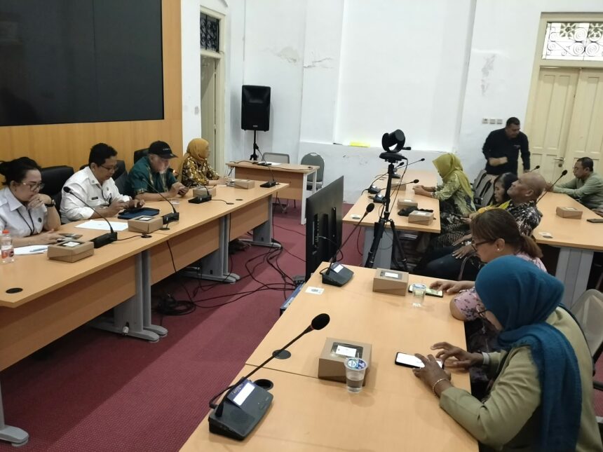 People sit at a long conference table with microphones and boxed favors during a meeting; a camera on a tripod is set up in the center of the room.