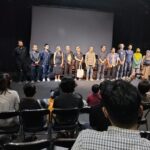 A line of diverse performers standing on a stage in front of a seated audience in a theater.