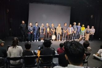 A line of diverse performers standing on a stage in front of a seated audience in a theater.