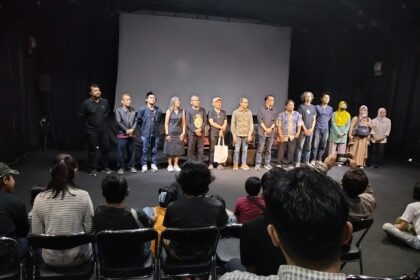 A line of diverse performers standing on a stage in front of a seated audience in a theater.