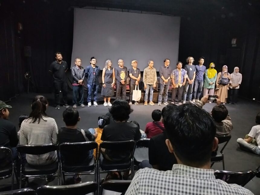 A line of diverse performers standing on a stage in front of a seated audience in a theater.
