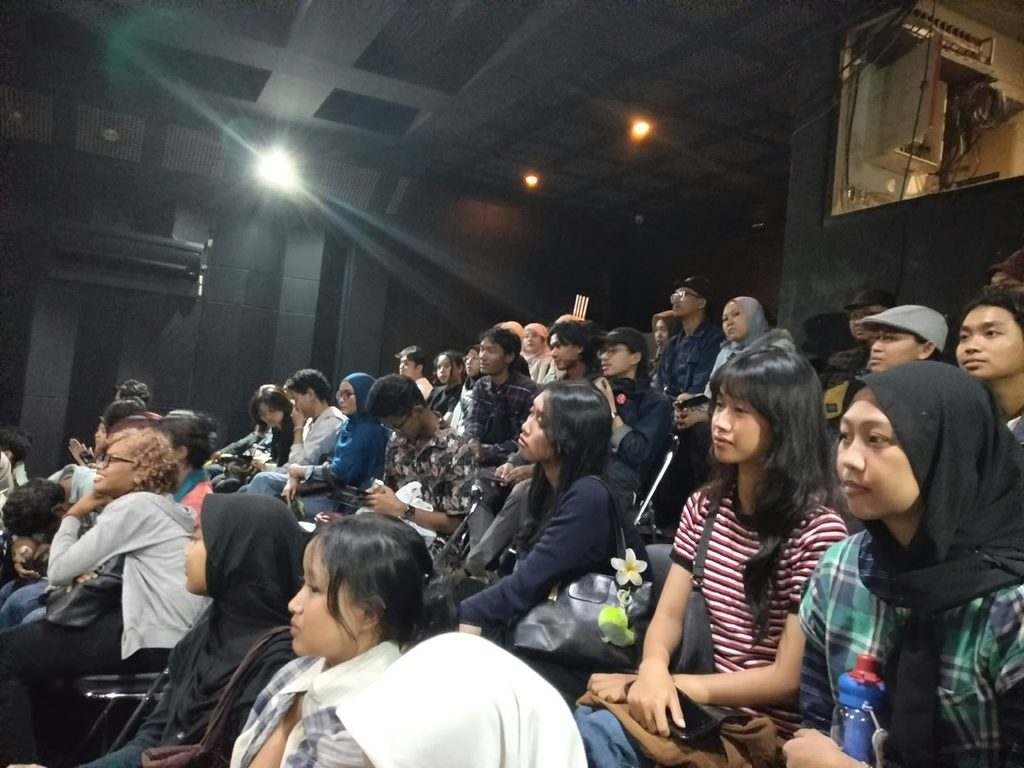 Diverse audience seated in a dim theatre, attentively watching a performance or talk.
