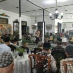 Community gathering in a decorated hall with a speaker at a microphone addressing seated guests in traditional attire.