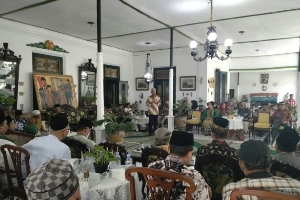 Community gathering in a decorated hall with a speaker at a microphone addressing seated guests in traditional attire.