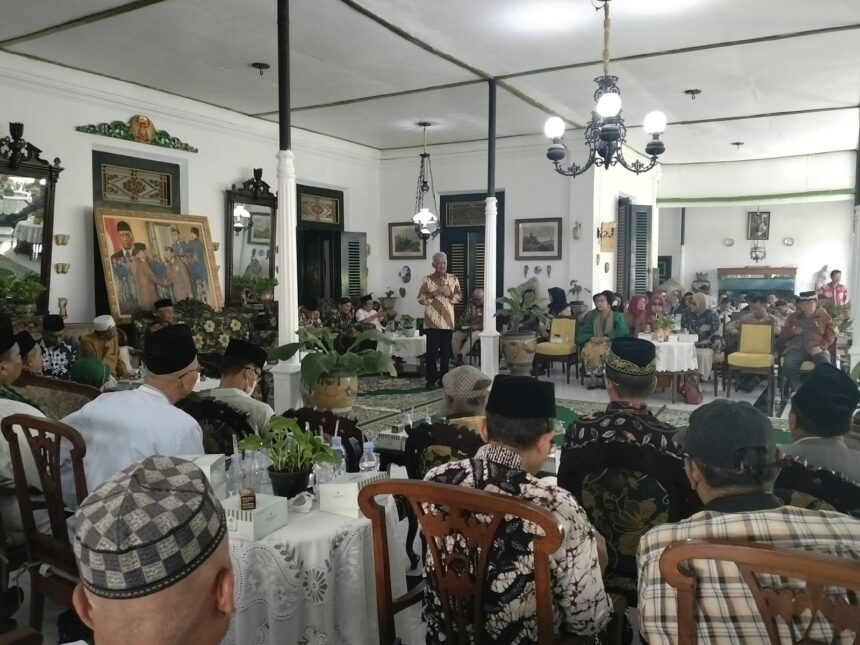 Community gathering in a decorated hall with a speaker at a microphone addressing seated guests in traditional attire.