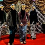 Four girls wearing hijabs perform on a red-carpet stage, each in patterned outfits and paper crown hats, with a guitar on the right backdrop behind them.