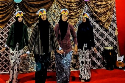 Four girls wearing hijabs perform on a red-carpet stage, each in patterned outfits and paper crown hats, with a guitar on the right backdrop behind them.