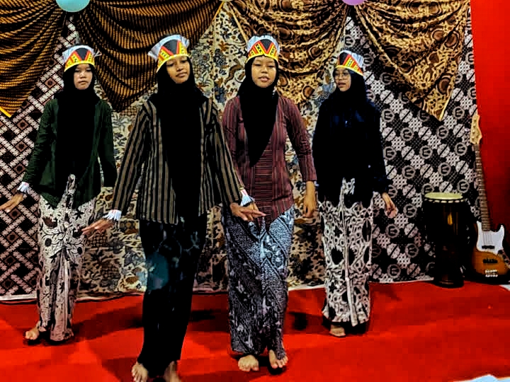 Four girls wearing hijabs perform on a red-carpet stage, each in patterned outfits and paper crown hats, with a guitar on the right backdrop behind them.