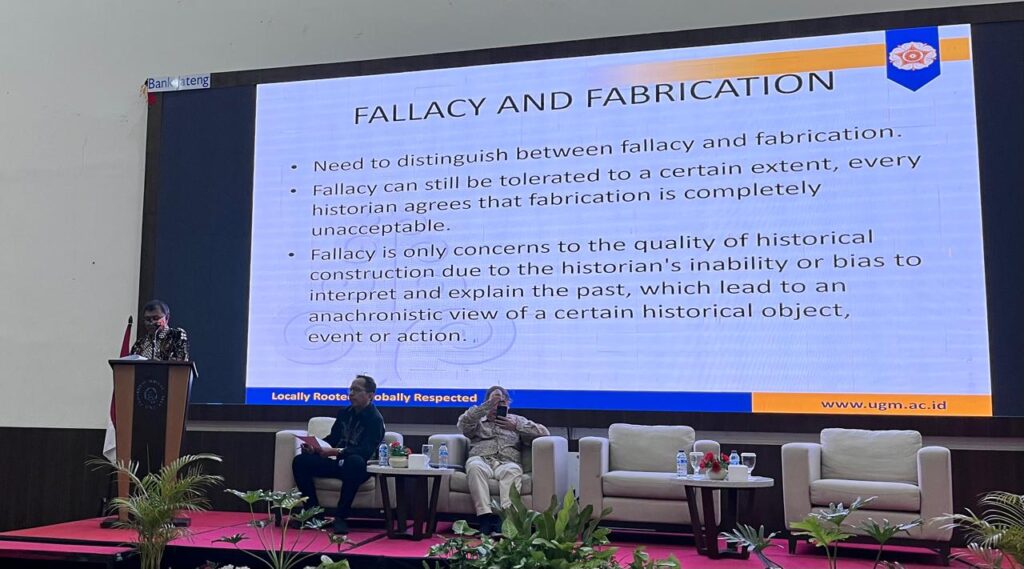 WhatsApp Image 2026-10-16 at 1226231 - Mabur.co Speaker at a podium on stage beside a large presentation slide reading 'Fallacy and Fabrication'; two panelists sit in white chairs, Indonesian flag visible on the left.