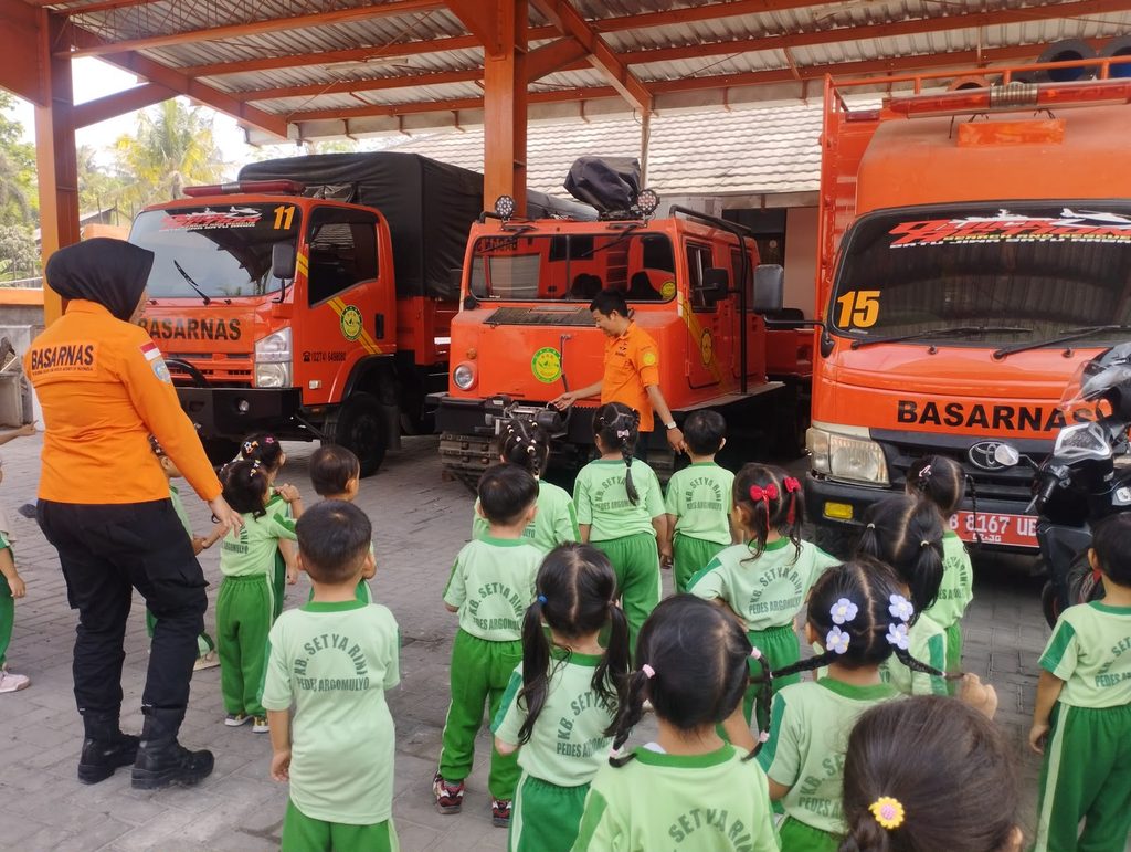 Group of young students in green uniforms listening to a rescue demo beside orange Basarnas emergency vehicles under a covered area.