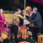 People on stage during a festive ceremony; a man in batik hands a wrapped gift to a smiling official while others in pink traditional attire look on.