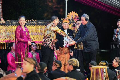 People on stage during a festive ceremony; a man in batik hands a wrapped gift to a smiling official while others in pink traditional attire look on.