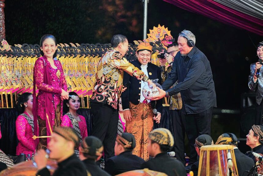 People on stage during a festive ceremony; a man in batik hands a wrapped gift to a smiling official while others in pink traditional attire look on.