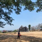 Stone temple ruin in an open park with a blue sky and trees framing the scene; a woman and child walk in the foreground.