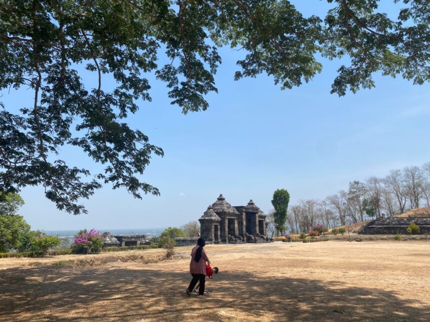 Stone temple ruin in an open park with a blue sky and trees framing the scene; a woman and child walk in the foreground.