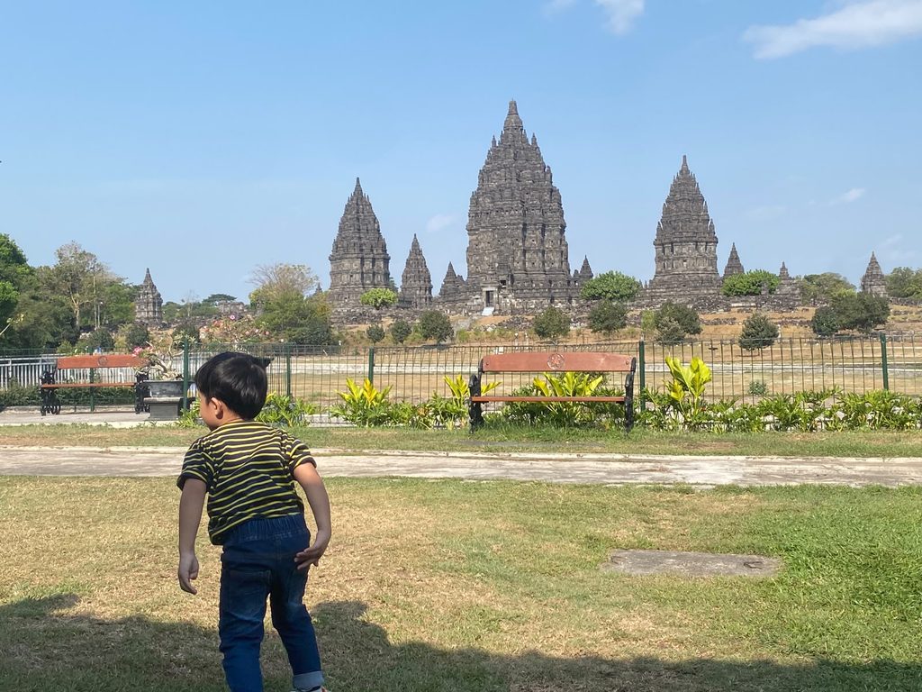 WhatsApp Image 2026-10-17 at 151735 - Mabur.co Young child walking in a park with ancient temple towers in the background under a clear blue sky.