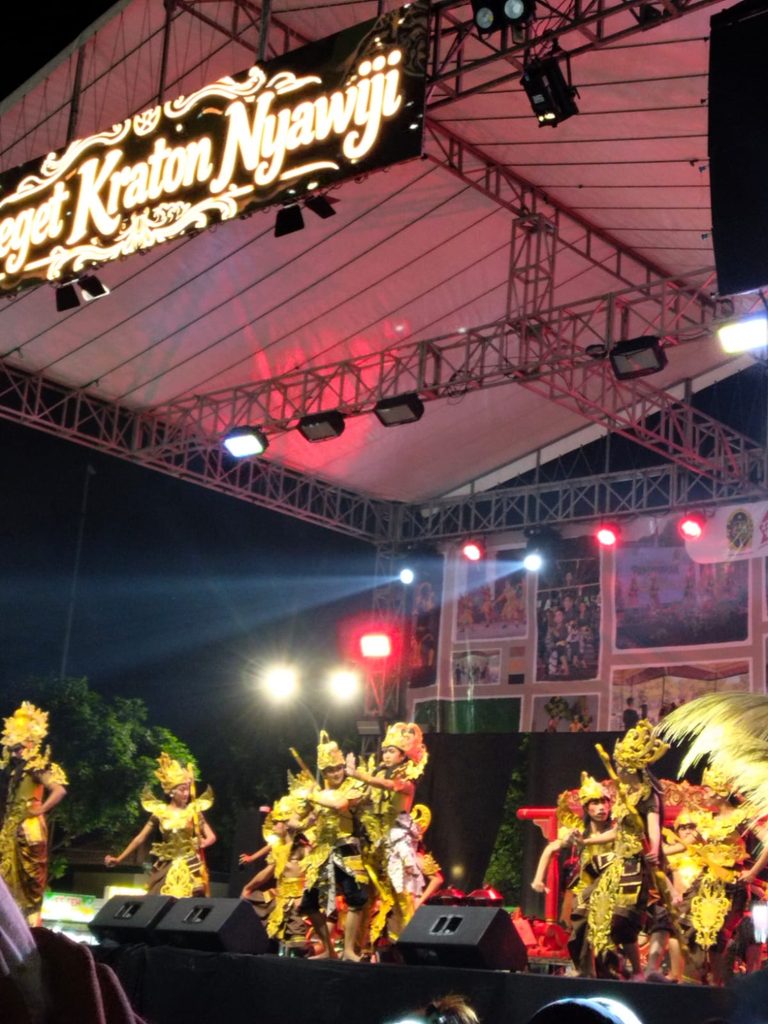Group of performers in elaborate gold costumes dancing on a lit stage under a decorative overhead sign at night, with a truss and stage lights above.