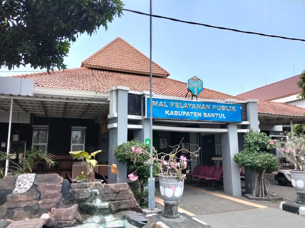 Front entrance of Bantul Regency Public Service Mall, blue sign reading 'MAL PELAYANAN PUBLIK KABUPATEN BANTUL' with a red-tiled roof and plants nearby.