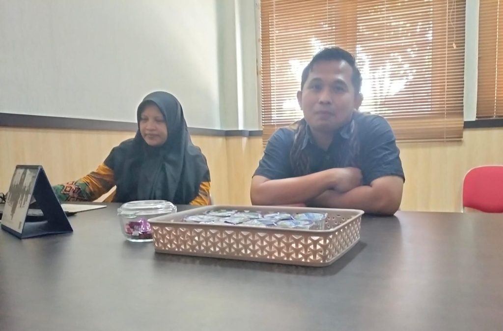 Two adults sit at a conference table: a woman in a hijab on the left and a man with folded arms on the right, in a room with blinds behind them.