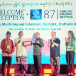 Five people on a stage clapping at the ICANN 87 Annual General Meeting welcome reception, with a bright screen displaying event text behind them and one person holding a plaque in the center.