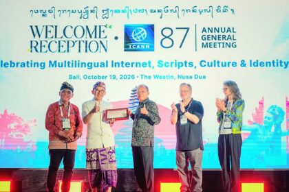 Five people on a stage clapping at the ICANN 87 Annual General Meeting welcome reception, with a bright screen displaying event text behind them and one person holding a plaque in the center.