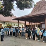 Group of people in blue shirts lined up in an outdoor courtyard, listening to an instructor at the front.