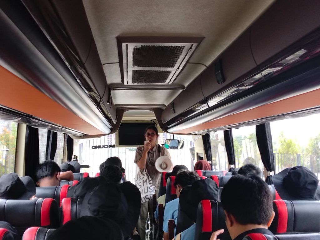 Bus interior with a guide standing at the front, speaking into a megaphone to seated passengers.