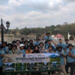 Group of tourists in matching light-blue shirts posing with a banner outdoors, with a decorative lamppost and hillside background.