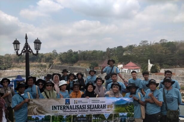 Group of tourists in matching light-blue shirts posing with a banner outdoors, with a decorative lamppost and hillside background.