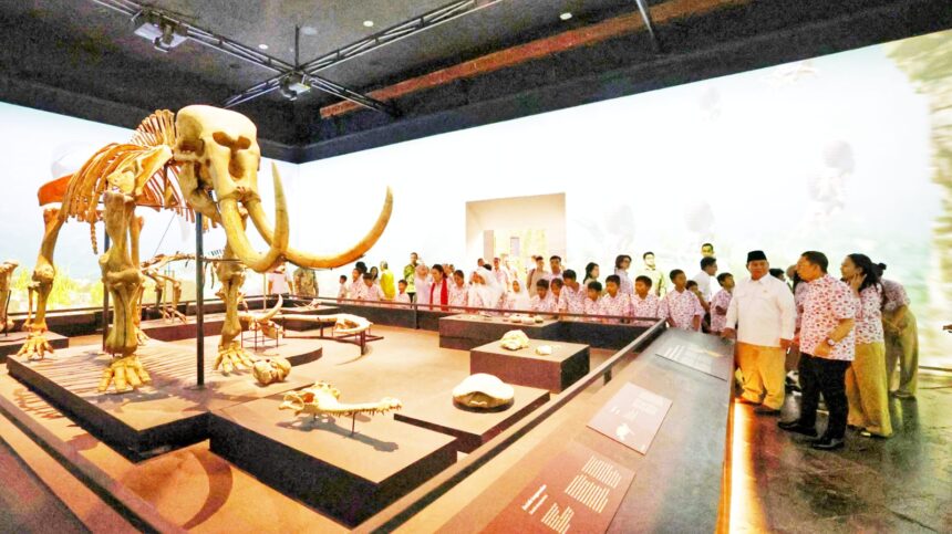 Large mammoth skeleton on a raised platform in a museum, with visitors gathered to view it.