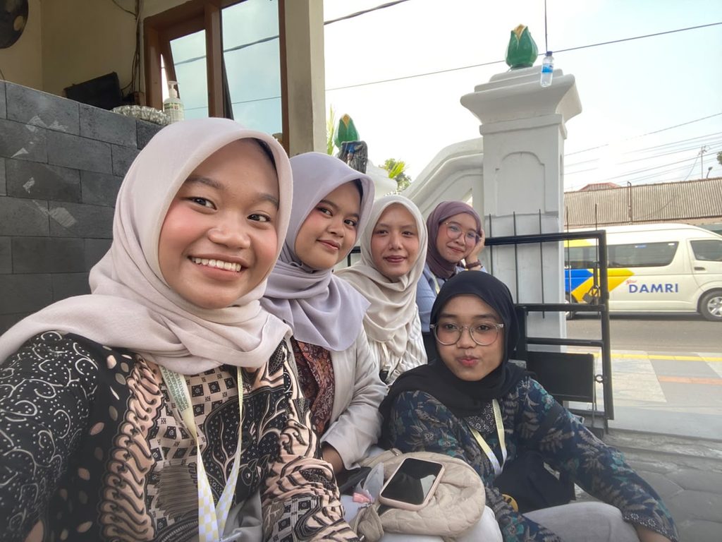 Five women wearing hijabs pose for a selfie outdoors near a white gate and a parked bus.