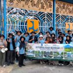 Group of people in blue shirts and hats pose with a large banner outdoors in front of an intricate blue gate backdrop.