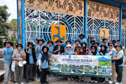 Group of people in blue shirts and hats pose with a large banner outdoors in front of an intricate blue gate backdrop.