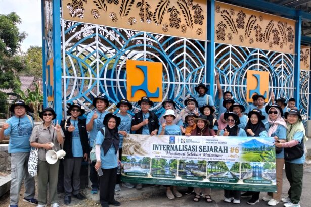 Group of people in blue shirts and hats pose with a large banner outdoors in front of an intricate blue gate backdrop.