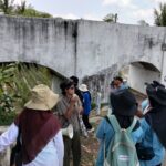 Tour group with backpacks listens to a guide near a weathered white concrete arch bridge in a tropical area.