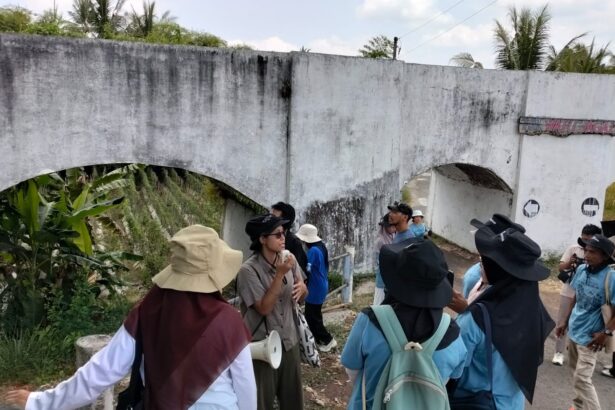 Tour group with backpacks listens to a guide near a weathered white concrete arch bridge in a tropical area.