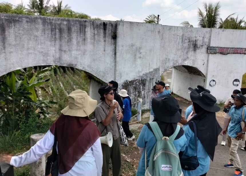 Tour group with backpacks listens to a guide near a weathered white concrete arch bridge in a tropical area.