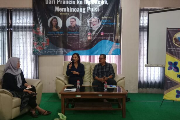 Panel discussion in a conference room with two people seated on a cream sofa and a woman on the left taking notes; a banner behind them advertises a poetry discussion event and logos are visible on the banner.