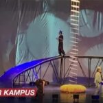 Live stage stunt show with performers on a tall ladder and curved bridge rig, blue lighting, and a yellow inflatable prop on stage; KABAR KAMPUS caption bottom left.