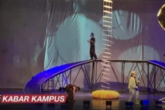 Live stage stunt show with performers on a tall ladder and curved bridge rig, blue lighting, and a yellow inflatable prop on stage; KABAR KAMPUS caption bottom left.