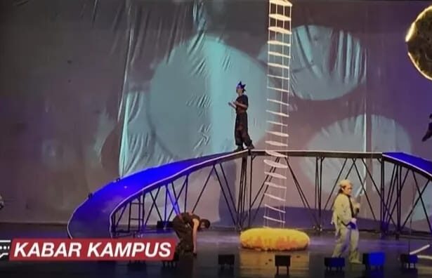 Live stage stunt show with performers on a tall ladder and curved bridge rig, blue lighting, and a yellow inflatable prop on stage; KABAR KAMPUS caption bottom left.