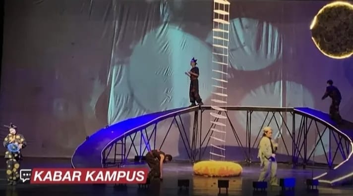 Live stage stunt show with performers on a tall ladder and curved bridge rig, blue lighting, and a yellow inflatable prop on stage; KABAR KAMPUS caption bottom left.