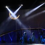 Stage performance with a glowing crescent moon prop overhead and blue-lit dancers along a curved, illuminated railing.