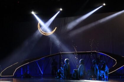 Stage performance with a glowing crescent moon prop overhead and blue-lit dancers along a curved, illuminated railing.