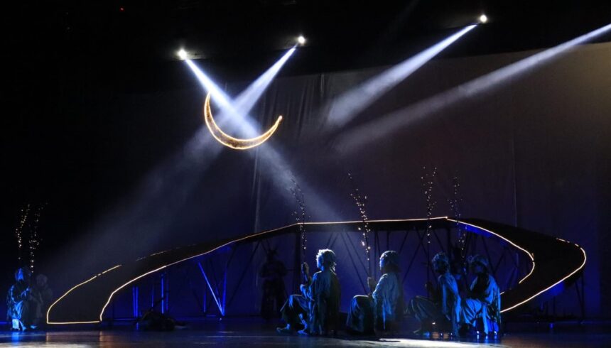 Stage performance with a glowing crescent moon prop overhead and blue-lit dancers along a curved, illuminated railing.