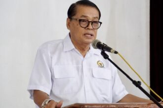 Man in a white short-sleeve shirt speaks at a wooden podium with a microphone in front of him.