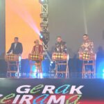 Six people stand behind wooden drums on a stage, performing a traditional drum ensemble under colorful stage lighting with a branded banner at the front.