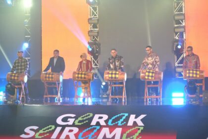 Six people stand behind wooden drums on a stage, performing a traditional drum ensemble under colorful stage lighting with a branded banner at the front.